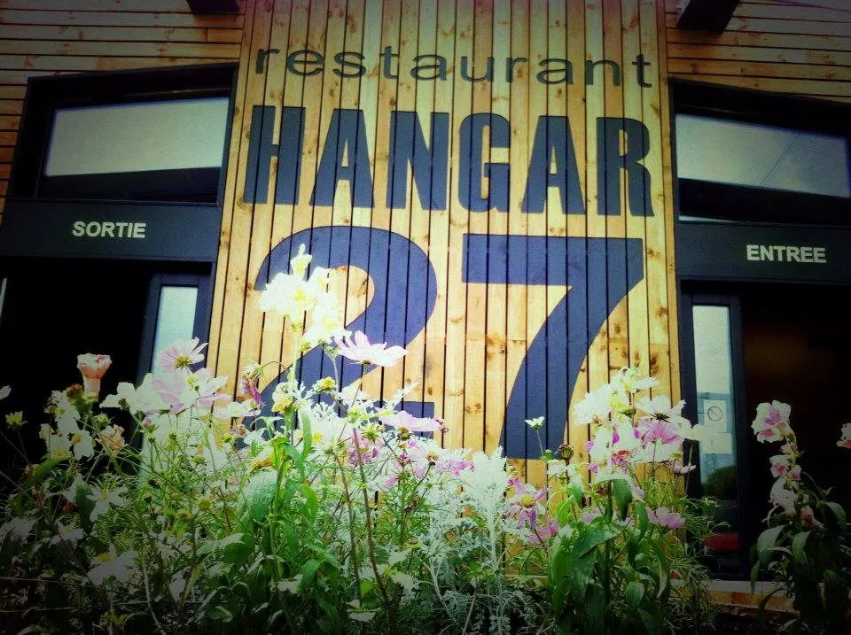 La fachada del Hangar 27: pared de madera, rótulo pintado y entrada florida